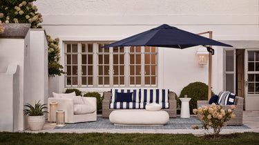 Outdoor patio setup with wicker sofa and armchairs, navy and white striped cushions, oval coffee table, large navy umbrella, and potted plants in front of a white house